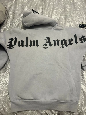 Grå Palm Angels hoodie med tryck - Snygg grå hoodie från Palm Angels med stort svart tryck på ryggen och mindre logga framtill. Tröjan har huva med svarta snören och en stor magficka. Perfekt för dig som gillar streetwear och vill sticka ut med en fet logga.