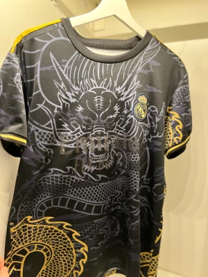 Real Madrid Ronaldo draktröja svart/guld - Unik Real Madrid fotbollströja med Ronaldo 7 på ryggen. Svart bas med detaljerat drakmönster i grått och guld, samt klubbmärke på bröstet. Gula ränder på axlar och ärmslut. Kortärmad och tillverkad i lätt polyester, perfekt för dig som vill sticka ut på planen.