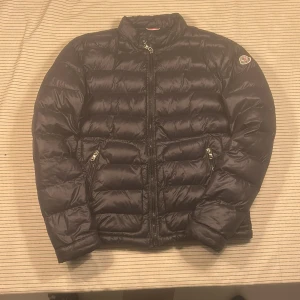 Svart Moncler pufferjacka herr - Svart pufferjacka från Moncler med klassisk logga på ärmen. Jackan har quiltad design, dragkedja framtill och två sidofickor med dragkedja. Insidan är röd med en cool serietidningsetikett. Perfekt för dig som vill ha en snygg och varm jacka till vintern