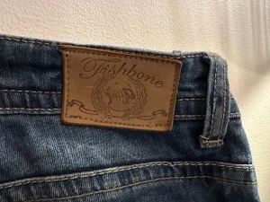 Lågmidjade blå Fishbone jeans med detaljer - Snygga blå jeans från Fishbone med slitningar och coola detaljer som dragkedja och små stjärnor vid fickan. Klassisk femficksmodell med låg midja och raka ben. Jeansen har kontrastsömmar och en unik knapp framtill. Perfekt för dig som gillar streetstil.