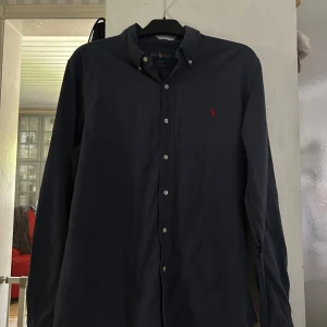 Mörkblå skjorta från Ralph Lauren - Klassisk mörkblå skjorta från Ralph Lauren med röd broderad logga på bröstet. Skjortan har knappar hela vägen, button-down krage och långa ärmar. Tillverkad i mjuk bomull och passar perfekt till jeans eller chinos.håll i vänster arm fast lätt att sy igen.passar även som M för det är slim fit 