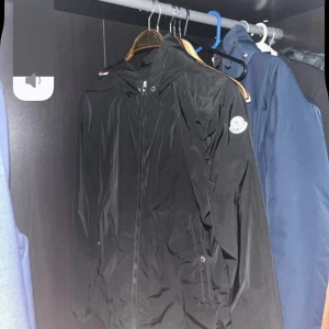 Svart vindjacka från Moncler - Svart vindjacka från Moncler med huva och dragkedja framtill. Jackan har Moncler-logga på vänster ärm och är tillverkad i lätt, glansig polyester. Perfekt för blåsiga dagar och har en clean, sportig look som funkar till streetwear.