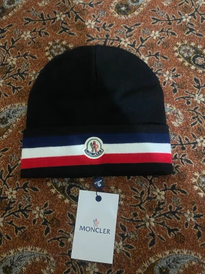 Svart Moncler mössa med ränder - Snygg svart mössa från Moncler med breda ränder i marinblått, vitt och rött längs kanten. Framtill sitter Monclers klassiska logotyp. Mössan är stickad och har en mjuk, skön känsla som passar perfekt när det är kallt ute.