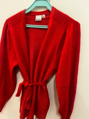 Röd kofta med knyt från ICHI - Snygg röd kofta från ICHI med långa ärmar och ribbade axelpartier. Koftan har ett bälte i midjan för knyt och är gjord i ett mjukt stickat material som känns skönt mot huden. Perfekt för att addera färg till din outfit. Använd en gång så färgen inte passade min smak 