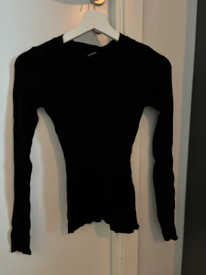 Svart ribbad långärmad topp Gina Tricot - Svart långärmad topp från Gina Tricot i slim fit med ribbad struktur. Rund halsringning och stretchigt material som sitter snyggt mot kroppen. Perfekt att styla med jeans eller kjol för en clean look.