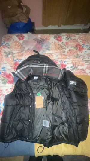 Svart Burberry pufferjacka med huva - Svart pufferjacka från Burberry med klassisk rutig huva i grått, vitt och rött. Jackan har dragkedja, knappar och två sidofickor med dragkedja. Märket 'Burberry London England' är broderat på vänster axel. Perfekt för kalla dagar och har en loose passform. Bara användt ett par gånger!