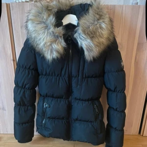 Svart pufferjacka med fuskpäls Hollies - Snygg svart pufferjacka från Hollies med stor fluffig fuskpäls på huvan och leopardmönstrat foder. Jackan har dragkedja framtill, praktiska fickor med dragkedja och ribbade muddar vid ärmslut. Perfekt för kalla dagar och ger en cool vibe.