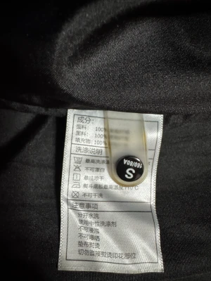 Svart lång pufferjacka från Nike - Svart pufferjacka från Nike med huva och två snedställda dragkedjefickor framtill. Jackan har en clean look med vitt Nike-logga på bröstet och långa ärmar. Perfekt för kalla dagar och har en längre modell som täcker bra. Materialet är polyester.