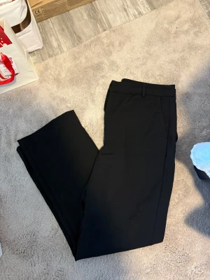 Svarta kostymbyxor med raka ben - Stilrena svarta kostymbyxor med raka, vida ben och klassiska hällor för bälte. Perfekta för en clean och dressad look. Byxorna har diskreta fickor och är sydda i ett mjukt tyg som ger en snygg passform.