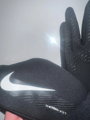 Nike svarta Therma-Fit handskar - Svarta Nike Therma-Fit handskar med vit swoosh-logga på ovansidan. Slimmad passform med mönstrad greppyta på insidan av fingrarna och tumme, samt touchfunktion på fingertopparna. Perfekta för träning utomhus när det är kallt.