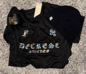 Decrese Outfit Paket – T-shirt Black Gloss + Bälte + Mössa. -  Decrese Sweden Outfit Paket – T-shirt + Bälte + Mössa.     Black Gloss Edition Svart T-shirt med holografiskt tryck som skiftar i färg för en sportig, Bälte: äkta läder, svart med silverfärgat spänne. Mössa: stilren svart med vit text.  Perfekt paket för en modern och stilren look.