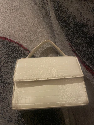 Vit handväska med krokodilmönster - Snygg vit handväska med krokodilmönstrad yta och stilren fyrkantig form. Väskan har ett praktiskt handtag upptill och stängs med magnetlås. Perfekt för dig som gillar trendiga accessoarer med lite edge och struktur.