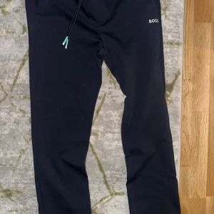 Svarta mjukisbyxor från BOSS, storlek S - Svarta mjukisbyxor från BOSS med logotyp på både fram- och baksidan. Byxorna har snörning i midjan och är tillverkade i mjukt bomullsmaterial. Passformen är normal och de har en enkel, stilren look som funkar till chill och häng.