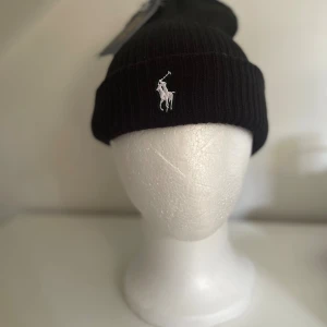 Svart ribbad mössa från Polo Ralph Lauren - Svart ribbstickad mössa från Polo Ralph Lauren med klassisk broderad logga i vitt framtill. Mössan har uppvikt kant och är tillverkad i mjukt stickat material som håller dig varm och snygg under kalla dagar. Tagen är ej kvar, fortfarande aldrig använd. 