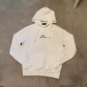 Vit hoodie från Polo Ralph Lauren XS - Vit hoodie från Polo Ralph Lauren i mjuk bomull. Tröjan har huva med snörning, broderad logga i svart på bröstet och en klassisk känguruficka framtill. Perfekt för chill dagar och enkel att matcha med jeans eller joggers.