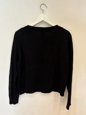 Svart stickad tröja från H&M - En enkel svart stickad tröja från H&M med rund hals och långa ärmar. Tröjan har ribbade muddar vid hals, ärmslut och nederkant. Perfekt för dig som gillar en clean och klassisk look.