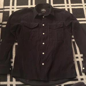 Svart manchester overshirt Mads Nørgaard - Svart overshirt från Mads Nørgaard i mjuk manchester med klassisk krage, vita knappar och två stora bröstfickor med lock. Långärmad och stilren design som ger en cool och avslappnad vibe. Perfekt att bära som lager över t-shirt eller hoodie.