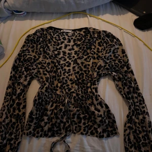 Leopardmönstrad blus från Zara - Säljer en snygg blus från Zara i leopardmönster med svarta och beige toner. Blusen har långärmade puffärmar, omlott-look och snörning i midjan för en cool siluett. Materialet är glansigt och ribbat vilket ger en extra touch. Perfekt för dig som gillar statement-plagg!