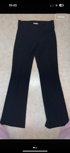 Svarta bootcut kostymbyxor från Nelly - Svarta kostymbyxor från Nelly i storlek 32. Byxorna har bootcut-ben och klassisk midja med hällor för bälte. Snyggt skräddad passform som ger en clean och stilren look. Perfekta för dig som gillar tidlös stil och vill ha ett par svarta byxor med lite vidd nedtill.