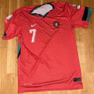 Portugal Ronaldo 7 fotbollströja Nike - Portugal landslagströja från Nike med nummer 7 och Ronaldo på ryggen. Röd med gröna detaljer, broderat landslagsmärke och Nike-logga. Tillverkad i lätt, ventilerande Dri-FIT-material. Perfekt för dig som älskar fotboll och Cristiano Ronaldo.