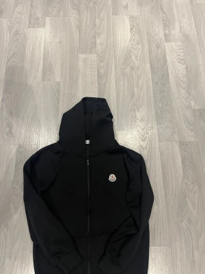 Svart Moncler hoodie med dragkedja - Svart hoodie från Moncler med klassisk logga på bröstet. Tröjan har huva, hel dragkedja framtill och ribbade muddar vid ärmslut. Materialet är mjuk bomull som känns skönt mot huden. Perfekt för dig som gillar stilrena och exklusiva märken.