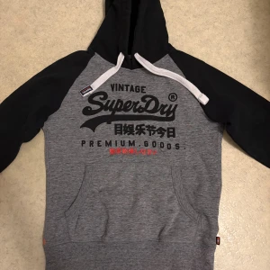 Grå och svart Superdry hoodie M - Snygg hoodie från Superdry i grått med svarta ärmar och huva. Stor logotyp och japansk text framtill, vita snören och känguruficka. Insidan är mjuk och fleecefodrad för extra komfort. Perfekt för chill dagar och streetstyle.