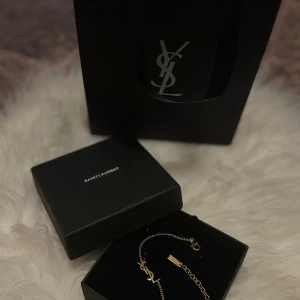 Saint Laurent guldarmband med YSL-logga - Exklusivt guldarmband från Saint Laurent med den ikoniska YSL-loggan i metall. Kedjan är tunn och elegant med en liten bricka där det står 'Saint Laurent Paris'. Perfekt accessoar för dig som gillar lyxiga detaljer och stilren design.