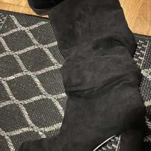 Svarta höga Boots/stövletter mocka - Säljer ett par svarta höga Boots med klack i mjuk mocka. Dem har en stilren och enkel design med rund tå och platt sula, perfekta för att lyfta din outfit. Passar dig som gillar klassiska och trendiga boots.Storleken är 39-42 Dem är helt nya Aldrig använt.Finns även samma men  i färgen Beige! Pris går att diskuteras!
