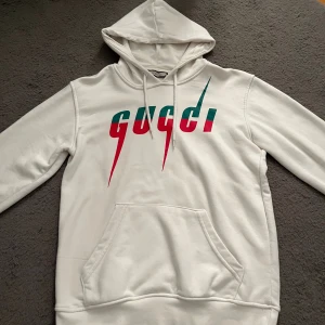 Vit hoodie  - Vit hoodie från GC med färgstarkt logotyp-tryck i grönt och rött framtill. Klassisk känguruficka, huva med snörning och långa ärmar. Mjuk bomullskänsla och relaxed fit – perfekt för dig som gillar streetstyle.