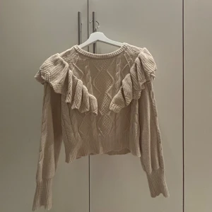 Stickad tröja  - Beige stickad tröja i storlek S med volang detaljer över axlarna och kabelstickat mönster framtill. Tröjan har rund halsringning, långa ärmar och ribbade muddar. Väldigt mysig och i bra skick.💕Skriv för mer!