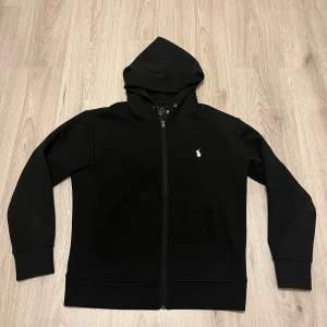 Svart hoodie från Polo Ralph Lauren - Svart hoodie från Polo Ralph Lauren med dragkedja framtill och klassisk vit broderad logga på bröstet. Tröjan har huva med snörning och två fickor framtill. Tillverkad i mjuk bomullsmix, perfekt för chill dagar eller när du vill ha en clean look.