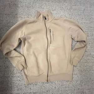 Äkta beige Sail Racing tröja.  - Beige fleecejacka från Sail Racing med hel dragkedja och hög krage. Jackan har en bröstficka med dragkedja och broderad logga på bröstet. Insidan är mjuk och fluffig, perfekt för kyliga dagar. Snygg och enkel design som funkar till allt. Har knappt används.         OBS pris kan diskuteras 
