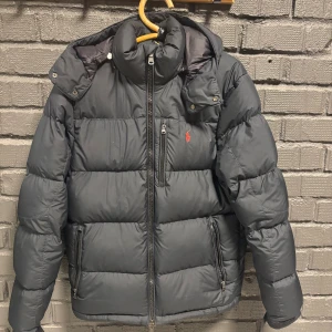 Svart pufferjacka från Polo Ralph Lauren - Svart pufferjacka från Polo Ralph Lauren med huva, dragkedja och flera fickor. Jackan har ett broderat rött Polo-logo på bröstet och är tillverkad i syntetmaterial. Perfekt för kalla dagar med sin tjocka dun fyllning och stilrena look. Kvitto finns digitalt.