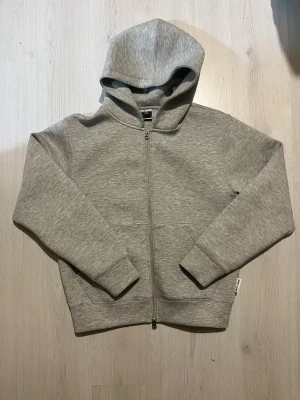 Grå hoodie från Jack & Jones Core - Tjena säljer nu den viral titkok hoodie från Jack a Jones med Core  dragkedja hela vägen och huva. Tröjan har ribbade muddar, två fickor fram och är tillverkad i mjuk bomullsmix. Perfekt för chill eller streetwear. Enkel och stilren design med diskret logga på sidan.