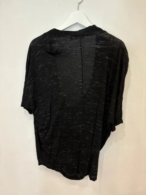 Svart draperad topp från Lindex - Svart topp från Lindex med draperad, djup v-ringning och korta ärmar. Tunt, lätt material med diskreta gråa ränder som ger en cool effekt. Oversized passform som faller snyggt och känns riktigt chill.