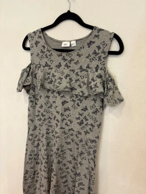 Grå kortklänning med fjärilsmönster från bpc - Supersöt grå kortklänning från bpc med svarta fjärilar över hela tyget. Klänningen har volangdetalj över bröstet och axlarna, samt ärmlös design med cut-out vid axlarna. Mjuk och skön bomullskvalitet, perfekt för varma dagar.