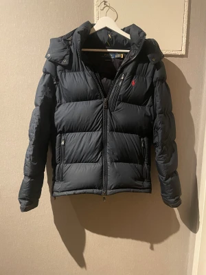 Svart pufferjacka Polo Ralph Lauren XS - Svart pufferjacka från Polo Ralph Lauren i storlek XS. Jackan har huva, två sidofickor med dragkedja och klassisk röd Polo-logga på bröstet. Materialet är polyester och jackan har en quiltad design med dragkedja framtill. Perfekt för kalla dagar. Köpte denna för 5200 på hanzenz 
