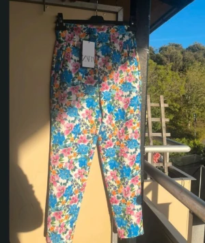 Blommiga croppade byxor från Zara - Färgglada croppade byxor från Zara med blommigt mönster i blått, rosa, orange och grönt på vit botten. Byxorna har hög midja och raka ben, perfekta för dig som vill sticka ut med en cool och somrig vibe. Prislappen är kvar 