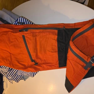 Orange täckbyxor från Fjällräven - Snygga orange täckbyxor från Fjällräven med svarta förstärkningar på knän och benslut. Byxorna har flera praktiska dragkedjefickor och bälteshällor. Perfekta för utomhusaktiviteter och äventyr. Modellen är för kvinnor och har en cool outdoor-look. Aldrig använda 