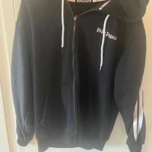 Svart hoodie Palm Angels 3XL - Svart hoodie från Palm Angels i storlek 3XL men sitter mer som M med vita snören och dragkedja framtill. Snygga vita ränder längs båda ärmarna och klassisk logga på bröstet. Hoodien har huva och stora fickor framtill, perfekt för en chill streetwear-look.
