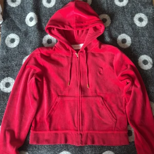 Juicy Couture hoodie - Säljer en röd hoodie från Juicy Couture med mjukt fleecematerial, huva med snörning och praktisk dragkedja framtill. Tröjan har långa ärmar och fickor på magen, perfekt för chill dagar. Liten broderad logga på bröstet för extra stil.