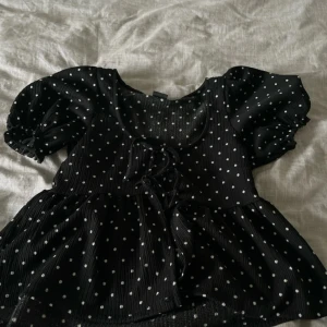 Svart prickig blus från Gina Tricot - Supersöt svart blus från Gina Tricot med vita prickar, puffiga korta ärmar och knytning framtill. Blusen har en lätt rynkad, flowy passform och ribbad struktur som ger en trendig look. Perfekt för dig som gillar lekfulla detaljer och vill sticka ut.