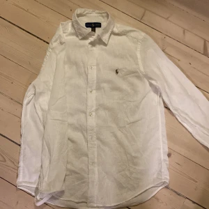 Vit linneskjorta från Ralph Lauren XL - Klassisk vit skjorta från Ralph Lauren i luftig linne, med broderad logga på bröstet. Skjortan har lång ärm, knappar framtill och en tidlös krage. Perfekt för en clean och stilren look.