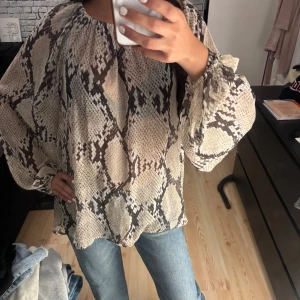 Blus - Säljer en lös blus från H&M med beige och brunt ormmönster. Blusen har långa, vida ärmar med resår vid handlederna och en rundad halsring. Knappt använd och som ny