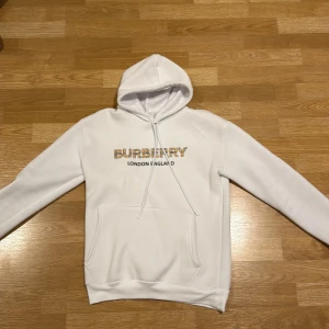Vit hoodie Burberry London England S - Vit hoodie från Burberry med klassiskt rutigt Burberry-tryck på bröstet och texten 'London England' under. Tröjan har huva med snörning, magficka och mjukt fleecefoder på insidan. Perfekt för dig som gillar stilrena och ikoniska streetwear-plagg.
