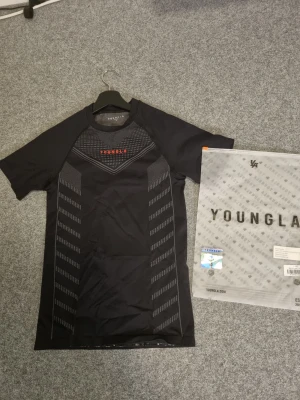 YoungLA compression - Svart t-shirt från trendiga märket YoungLA, grått grafiskt mönster framtill och röd logga på bröstet. Tillverkad i ett stretchigt syntetmaterial som passar perfekt för träning. 
