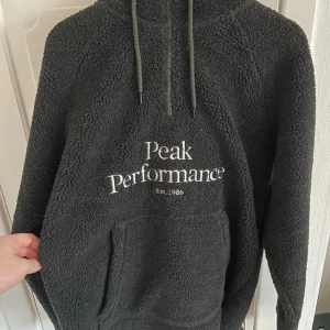 Svart fleece hoodie Peak Performance - Mysig svart hoodie från Peak Performance i storlek L. Tröjan är gjord i fluffig fleece med huva och snörning, samt en stor magficka framtill. Halv dragkedja upptill och broderad logga på bröstet. Perfekt för kalla dagar och chill.
