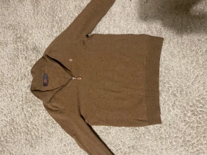 Brun polotröja Morris merinoull S - Snygg brun polotröja från Morris i 100% merinoull. Tröjan har lång ärm, ribbade muddar och en stilren half zip-dragkedja vid halsen. Perfekt för dig som gillar klassisk och clean stil med lite preppy vibe. 