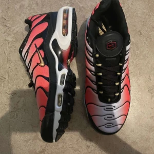 Nike Air Max Plus rosa och svart - Nike Air Max Plus sneakers med vågigt mönster i rosa, svart och vitt. Skorna har synlig Air Max-dämpning, svarta snören och guldigt swoosh på sidan. Ovandelen är i syntetmaterial med cool gradient-effekt och platt sula. Perfekt för dig som gillar att sticka ut.