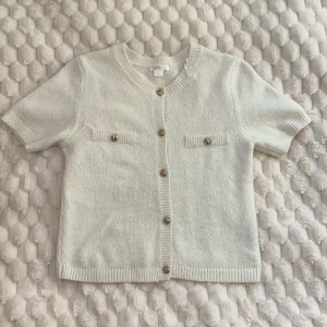 Vit kortärmad kofta från Gina Tricot - Supersöt vit kofta från Gina Tricot med korta ärmar och snygga guldfärgade knappar framtill. Koftan har två dekorativa fickor på bröstet och är stickad i mjukt material med ribbade kanter vid hals, ärmslut och nederkant. Perfekt för lager-på-lager och chill stil.